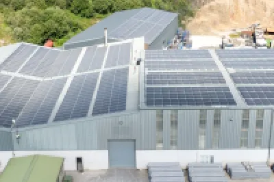 Industrial facility with large solar panels on the roof, surrounded by parked vehicles and storage materials.