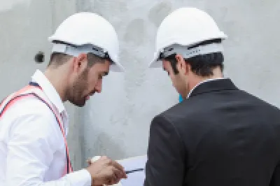 Two people wearing white safety helmets reviewing documents at a construction site in front of a concrete wall.