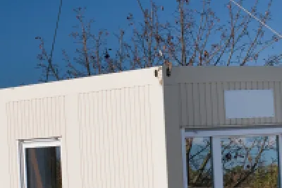 Prefabricated modular unit being lifted by a crane onto a site, with two workers in safety gear guiding it into place.