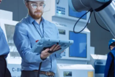 Two engineers in hard hats use a tablet to program a robotic arm on a factory floor.