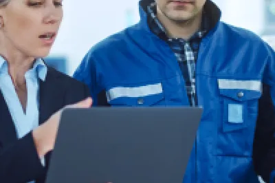 Two people wearing hard hats in an industrial setting, one in a business suit holding a laptop and the other in a blue work jacket, reviewing information together.