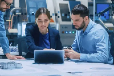 Three people in a modern office sitting around a table with a laptop, reviewing documents and technical drawings, with machinery and monitors visible in the background.