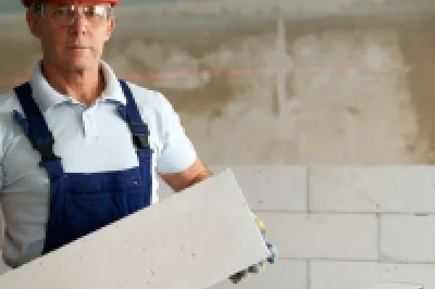 Construction worker in a red hard hat and overalls holding a large white block indoors, with a step ladder and unfinished wall in the background.