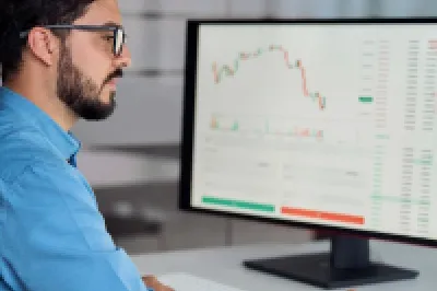 Person in a blue shirt working at a desk with a laptop and large monitor displaying financial charts and data.