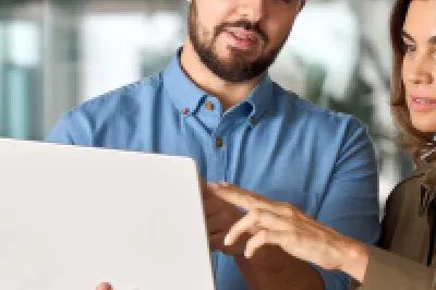 Two people standing in an office, holding a laptop and a tablet, discussing something on the laptop screen.