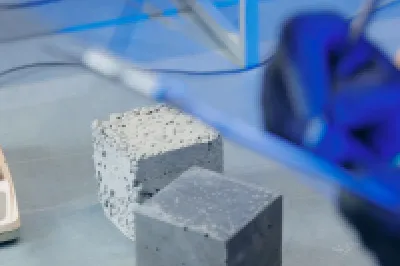 Concrete test cubes on a table, with one cube placed on a digital scale and a person wearing gloves holding a clipboard in the foreground.