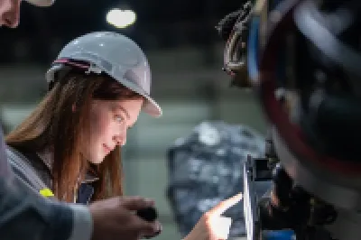 Two people wearing safety helmets and reflective clothing inspecting industrial machinery, with one person pointing at a control panel.