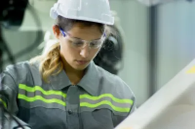 Person wearing a hard hat, safety glasses, and protective clothing operating equipment in an industrial setting.