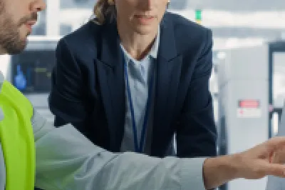 Engineer in a safety vest and hard hat pointing at a computer screen while discussing data with a colleague in a hard hat inside an industrial facility.