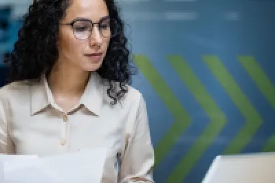 Person with curly hair and glasses sitting at a desk, holding papers and looking at a laptop screen in an office setting.