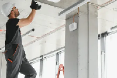 Person wearing a hard hat and overalls standing on a ladder and installing or servicing a ceiling-mounted air conditioning unit in a modern building.