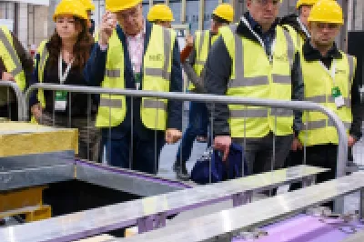 Participants in high-visibility vests and helmets observing a construction demonstration inside a manufacturing facility.