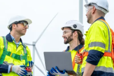 Three workers in safety gear discussing plans near a wind turbine.