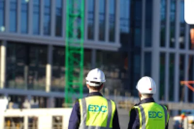 Two people wearing hard hats and high-visibility vests with the EDC logo, observing a modern glass building under construction.