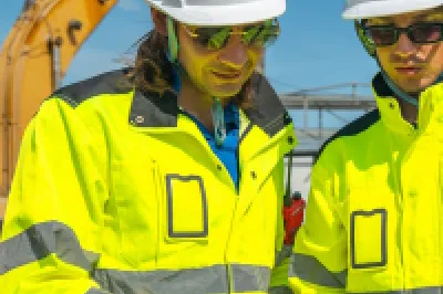 Two construction workers in hard hats and high-vis jackets reviewing plans at a building site.