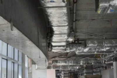 Construction site interior showing exposed ceiling with pipes and ductwork, and unfinished concrete walls.