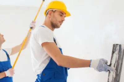 Two workers in yellow hard hats and blue overalls plastering a white interior wall.