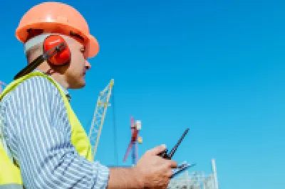 Construction worker in safety vest and helmet using tablet at building site against blue sky.