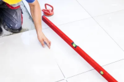 Construction worker in yellow hard hat laying white tile flooring with a red leveling tool.