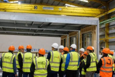 Group of workers wearing safety vests and hard hats gathered in an industrial warehouse facility.