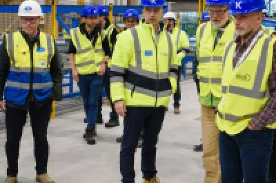 Group of people in hard hats and hi-vis vests touring a manufacturing facility with blue overhead crane.