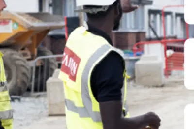 Two construction workers in safety vests and hard hats at a building site, with company logos visible.