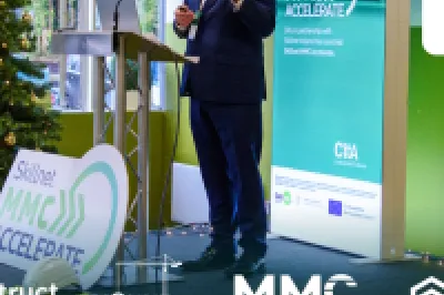 A person speaking at a podium with a Christmas tree and event sponsor banners in the background.