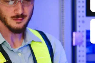 Worker in blue hard hat and safety vest using tablet near electrical control panel.