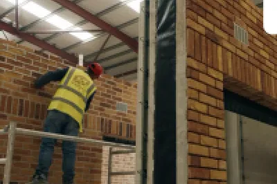 Worker in yellow vest on ladder inspecting large brick-effect modular structure in industrial warehouse.