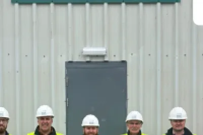 Six workers in hi-vis vests and hard hats stand in front of an MMC building with Mount Lucas logo.