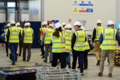Group of workers in yellow safety vests touring a large industrial warehouse facility.