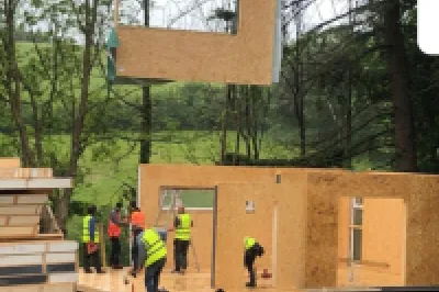 A crane lifts a modular wooden wall panel onto a construction site, with workers guiding it into place.