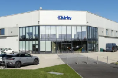 Modern industrial building with "Eleby" signage, cars parked outside under a cloudy sky.
