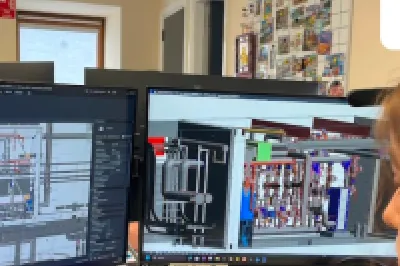 Woman working at a dual-monitor workstation displaying technical CAD drawings.