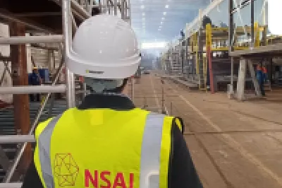 Worker in NSAI yellow vest and hard hat inspecting a large industrial facility.