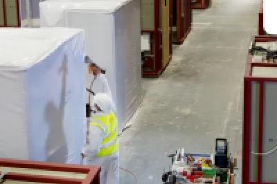 Factory workers preparing prefabricated construction modules inside a large manufacturing facility. Numerous partially assembled modules are arranged throughout the factory floor.
