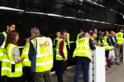 Group of visitors in yellow safety vests touring an industrial facility with large black equipment.