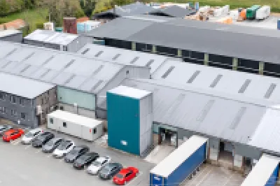 Aerial view of a large industrial warehouse complex with trucks and vehicles parked outside, surrounded by green countryside.