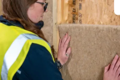 Woman in yellow safety vest installing mineral wool insulation panels in a wooden wall frame.