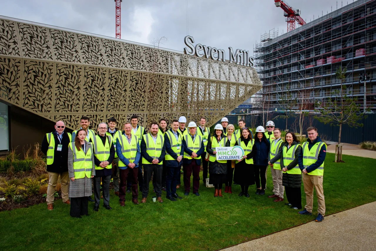 Group of workers in yellow safety vests standing on grass in front of a construction site.
