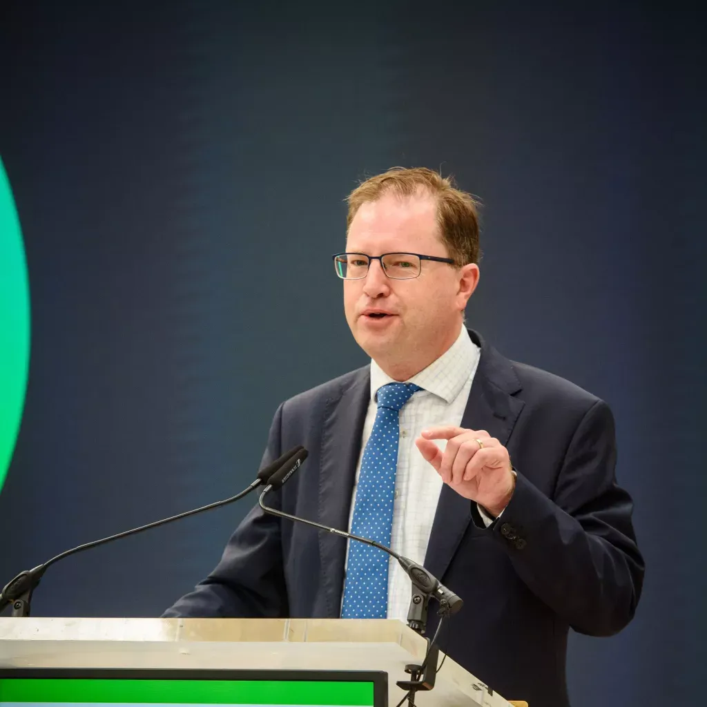 A person in glasses and suit speaking at a podium with a green accent on stage.