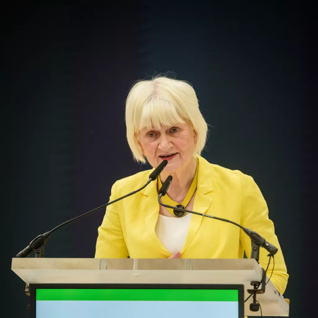Woman in bright yellow jacket speaking at a podium with microphone against dark background.