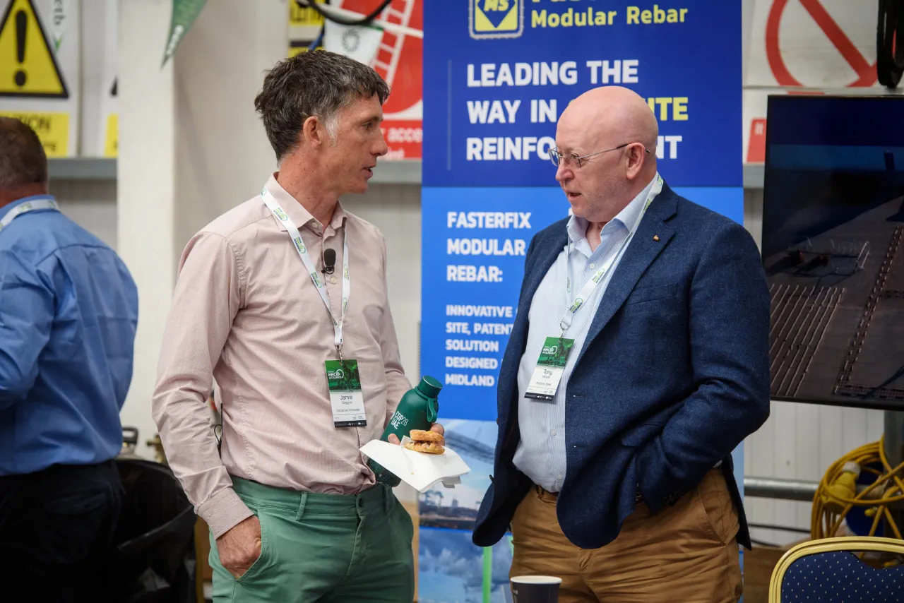 Two people in conversation at an event, one wearing a light shirt and the other in a dark blazer.