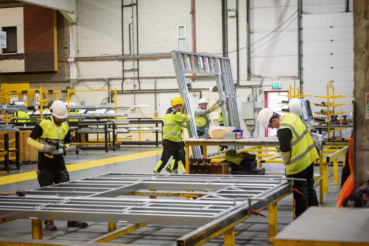 Construction workers in yellow high-visibility vests working on a manufacturing floor with metal frames.