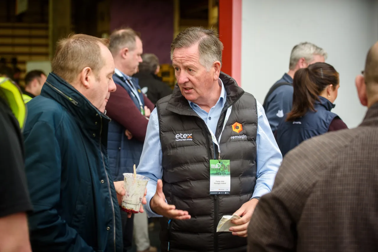 Two men in conversation at a business event, one wearing a dark jacket and the other a gray vest.