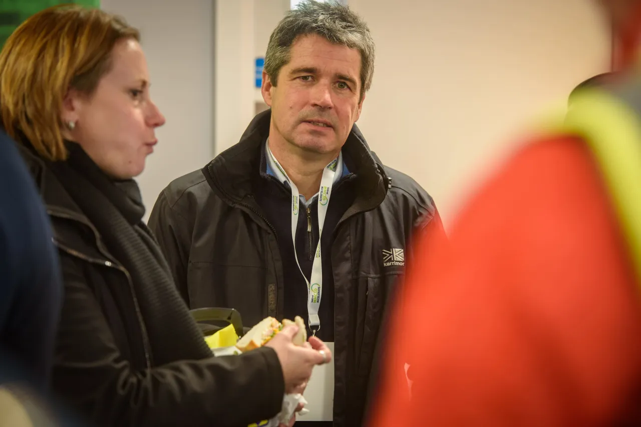 Two people in conversation with plates of food, one with gray hair wearing a dark jacket.
