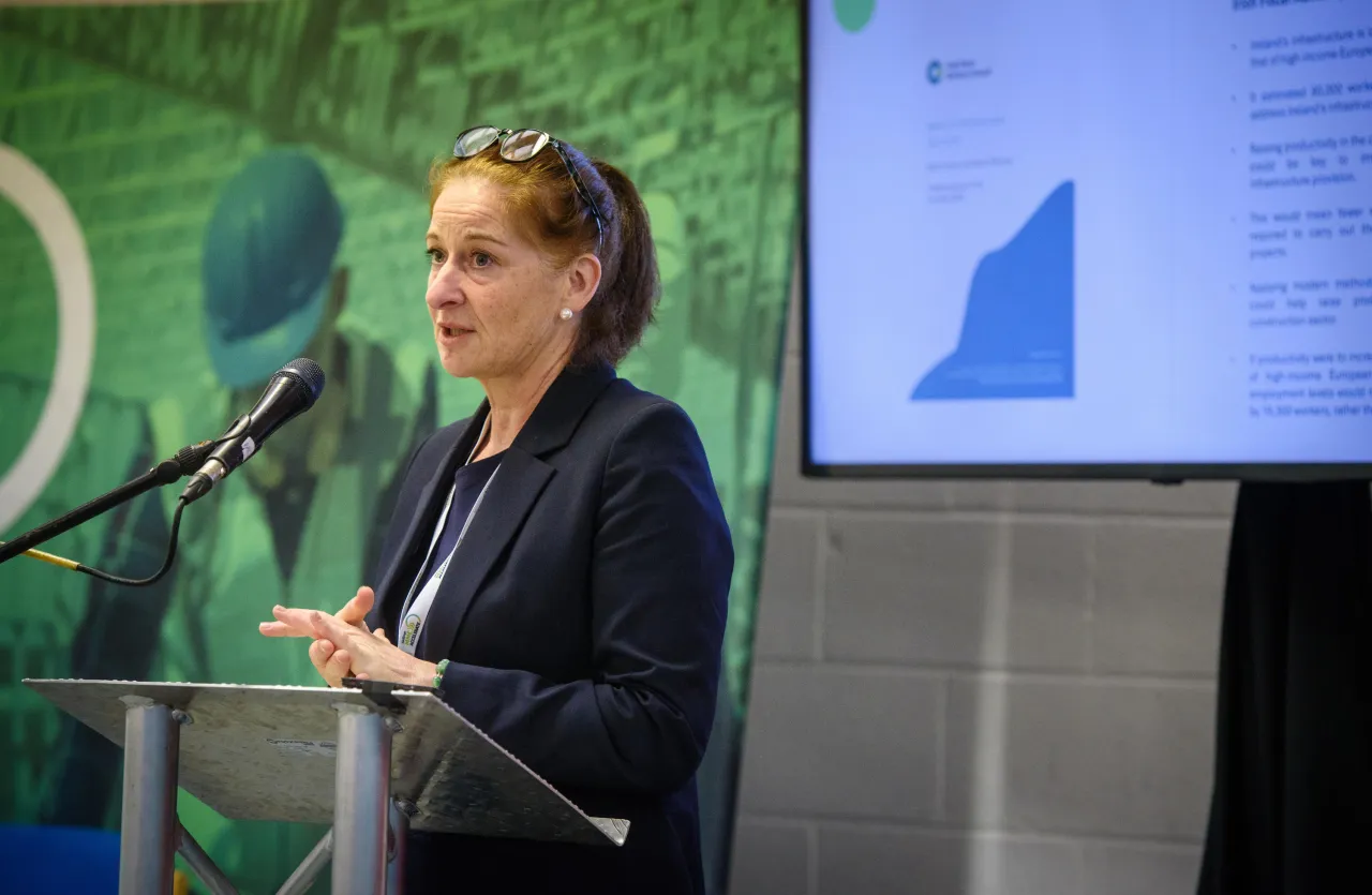 A person speaking at a podium with a green backdrop, presentation screen visible to the right.