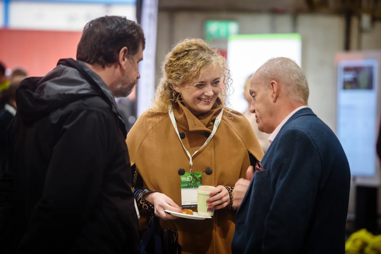Three people standing together in conversation, one person holding a drink in a tan coat.