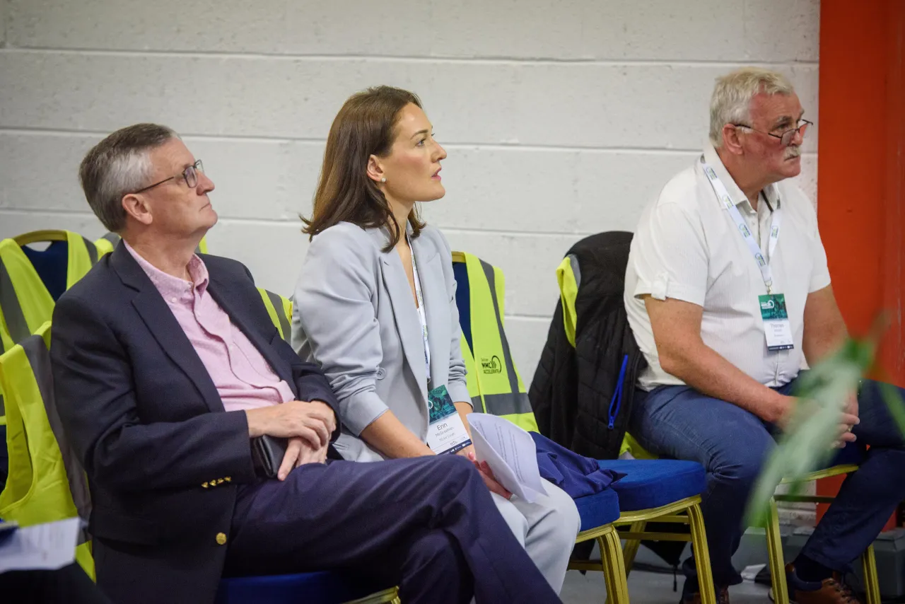 Three people seated in blue chairs at an event, two wearing safety vests.