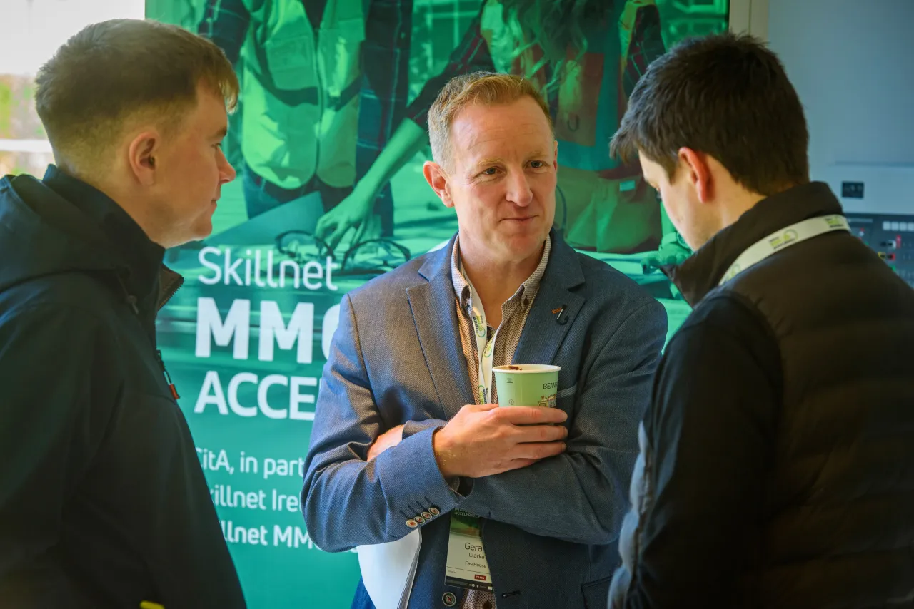Three people in conversation at a conference with green promotional banners in the background.